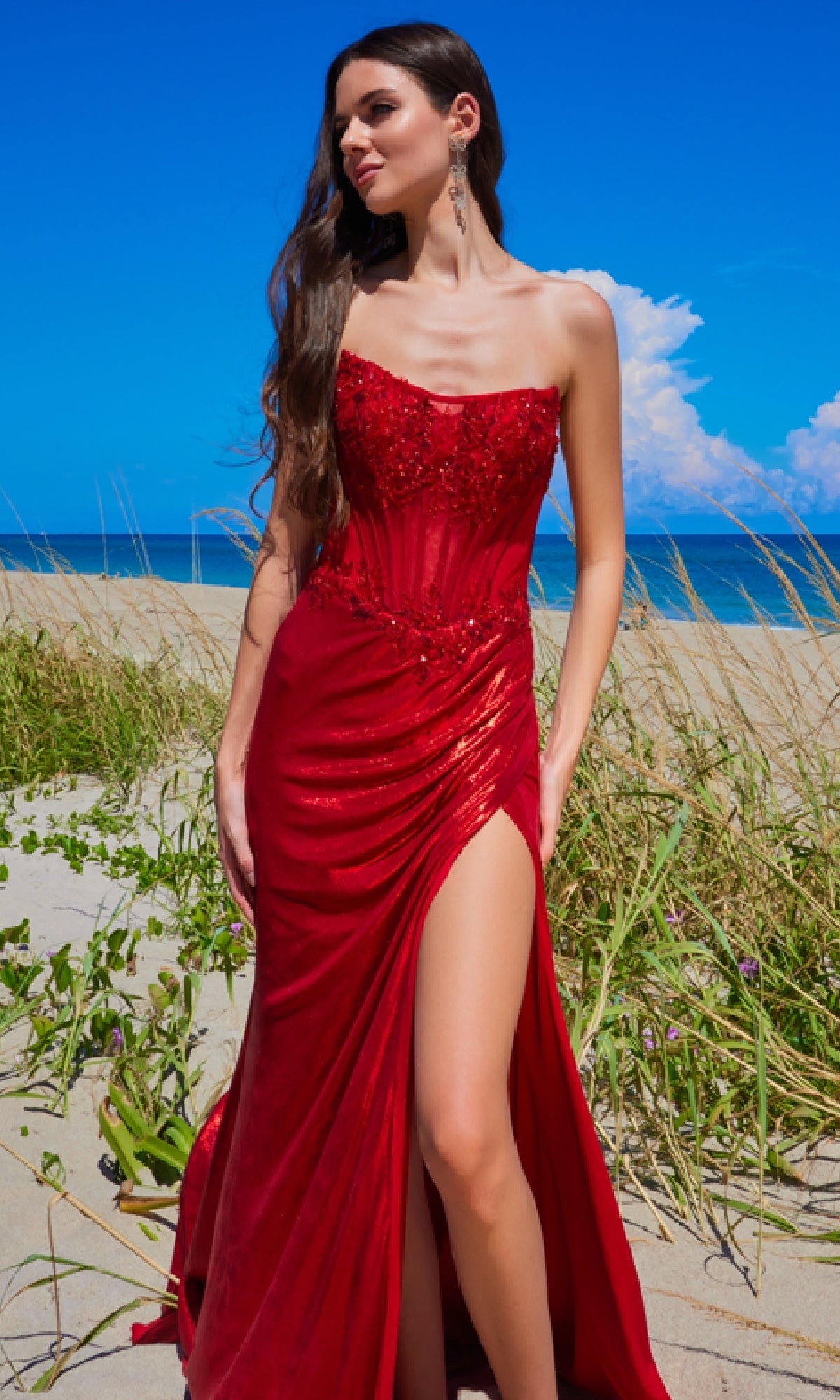 A woman wearing the Amelia Couture 9055 Strapless Long Prom Dress with a corset-style bodice stands on a sandy beach with green grass, beneath a clear blue sky.